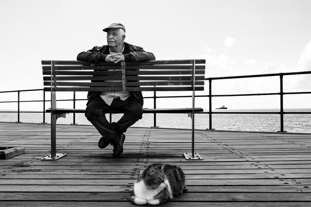 A black and white photo of an elder man sitting on a bench with his hands crossed. In front of him there is a cat sitting with its legs crossed